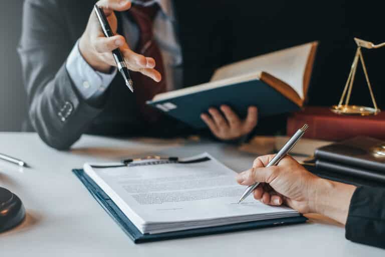 2 people discussing document on a clipboard, one hand holding a pen, other person holding a law book