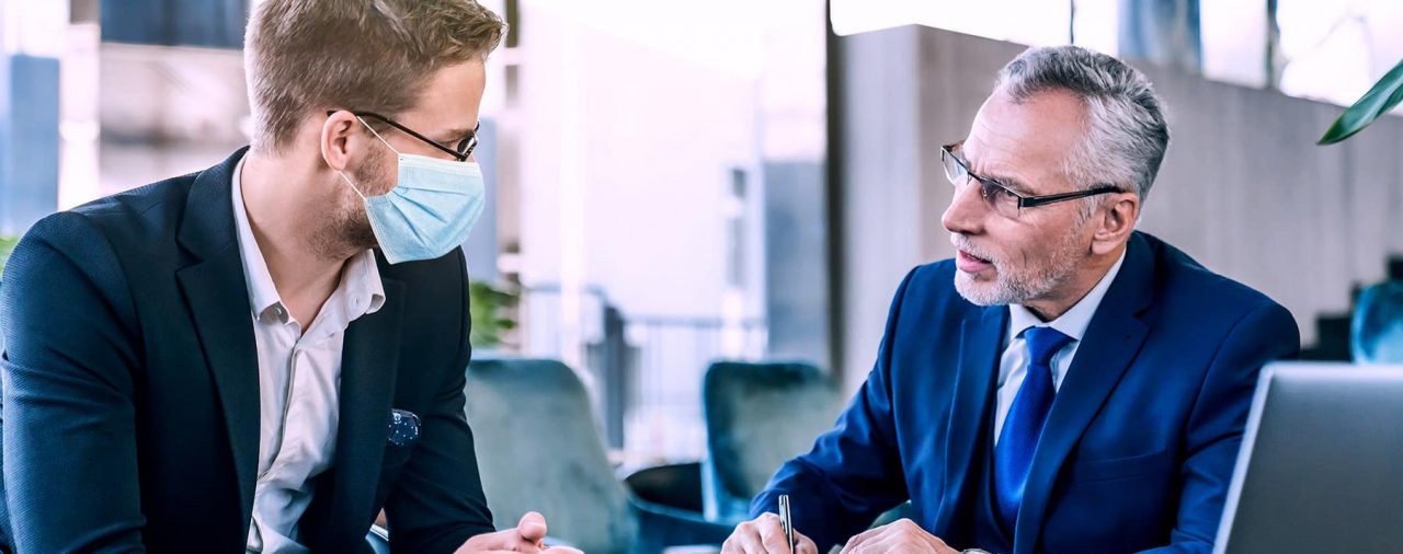 Business men at desk, one with mask, having discussion