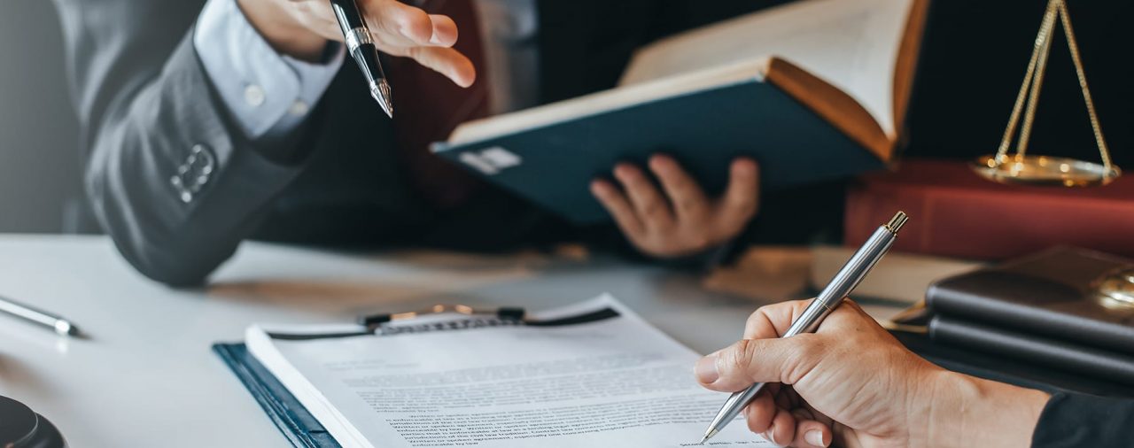 2 people discussing document on a clipboard, one hand holding a pen, other person holding a law book